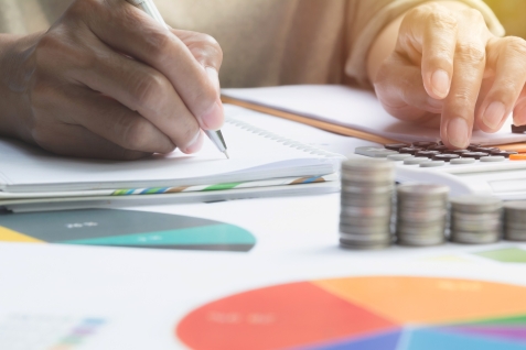 iemand typt op calculator en schrijft mee, op voorgrond pie-charts en stapels muntgeld