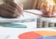 iemand typt op calculator en schrijft mee, op voorgrond pie-charts en stapels muntgeld